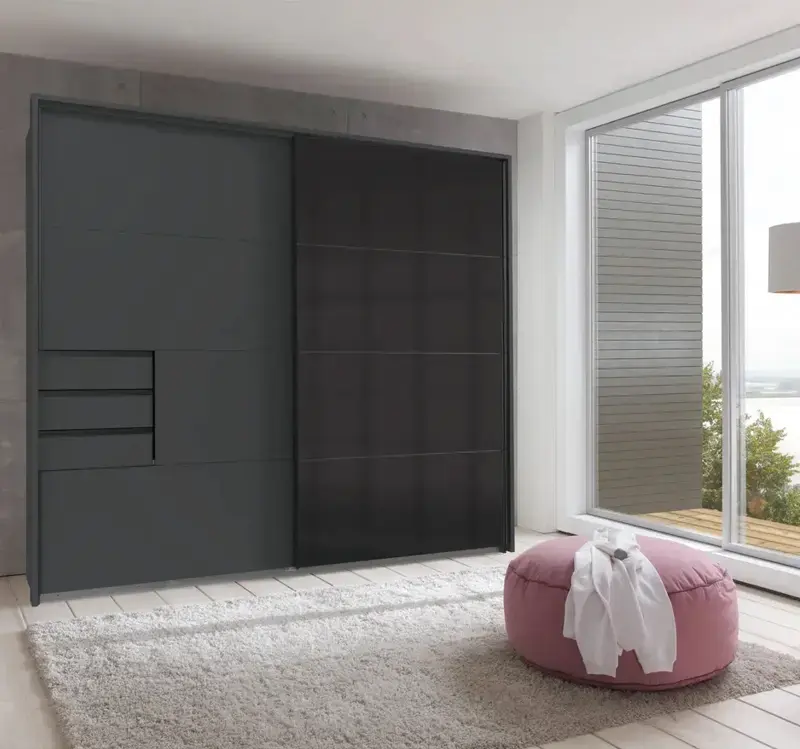 Schlafzimmer mit grossem Schiebetür Kleiderschrank in dunklem Grau links, rechts bodentiefe Fensterfront, davor rosa Pouf mit weissem Kleidungsstück auf Teppich.
